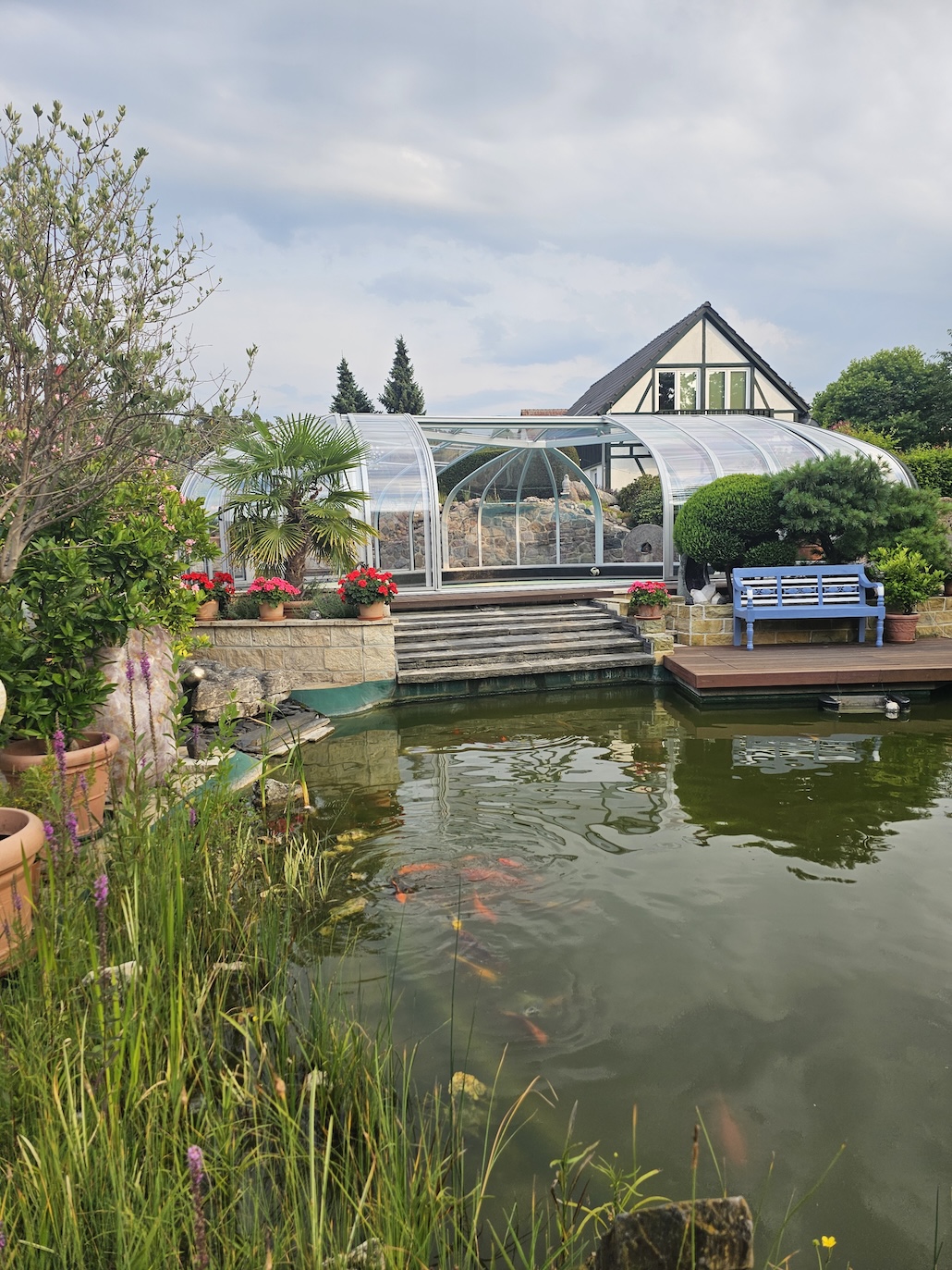 Begehbare Poolüberdachung in einem gepflegten Garten mit Teich, Steintreppe und Sitzbank